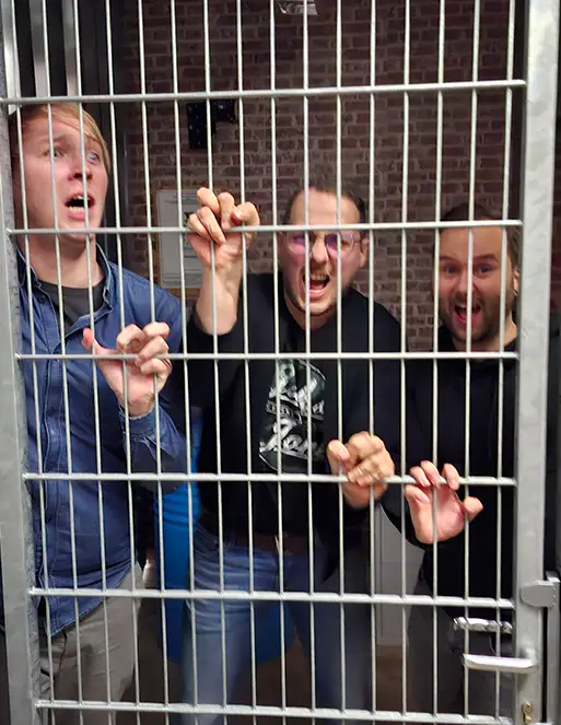 Auf dem Bild sind drei Männer. Sie stehen vor einer Gittertür und halten sich mit den Händen zwischen den Gittern fest.