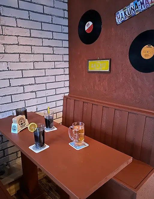 mystery_pub_01 Auf dem Bild ist eine Sitzbank mit einem Tisch. Auf dem Tisch stehen 4 Getränke. Auf der Wand hinter der Sitzbank sind zwei Schilder und zwei Schaltplatten aufgehangen.