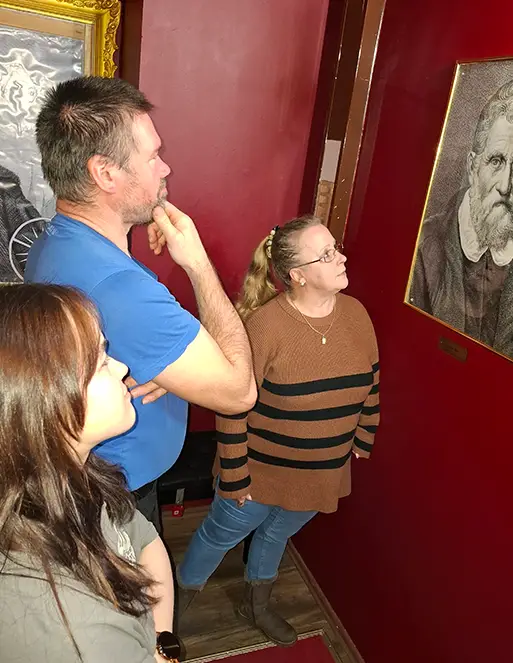 skriptorium_01 Ein Mann und zwei Frauen schauen sich ein Bild an, welches auf einer roten Wand hängt.