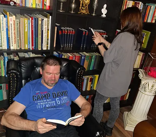 skriptorium_02 Ein Mann sitzt in einem schwarzen Sessel und ließt ein Buch. Hinter ihm ist ein Bücherregal an dem eine Frau steht und sich die Bücher anschaut.