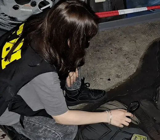 tatort_hamburg_02 Eine brünette Frau sitzt in der Hocke und hat eine Lupe in der Hand und beobachte damit den Boden.