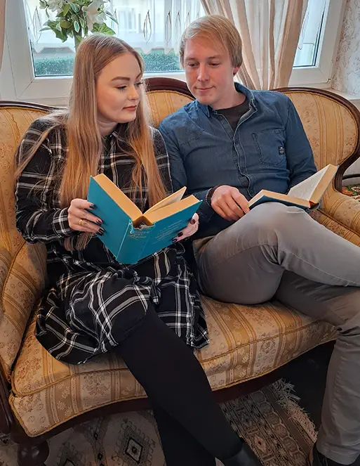 Ein Mann und eine Frau sitzen auf einem Sofa und schauen sich gegenseitig in das Buch von der anderen Person.