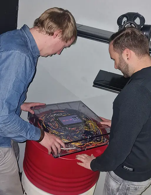 Auf dem Bild sind zwei Männer. Sie schauen beide auf einen Glaskasten mit vielen Kabel drin. Auf dem Glaskasten ist eine Zeit angezeigt.