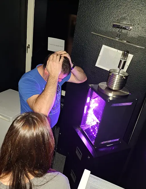 Auf dem Bild sind zwei Personen, die vor einem Gerät stehen, welches lila leuchtet. Der Mann schlägt sich die Hände auf den Kopf und die Frau neben ihm schaut den Mann an.